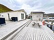 Holzterrasse am Skarsvåg Touristensenter in Nordnorwegen mit Blick auf den Hafen. Traditionelle Holzhäuser bieten gemütliche Unterkünfte, ideal für Angelurlaub. Fjordlandschaft im Hintergrund verstärkt das Naturerlebnis. Perfekte Kombination aus Ruhe und Abenteuer.