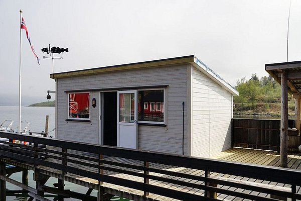Gemütliches Angelhäuschen am Wasser in Nordland, Norwegen. Weiße Holzhütte mit Terrasse, norwegische Flagge weht im Wind. Ideal für Angelurlaube und Entspannung in malerischer Umgebung.