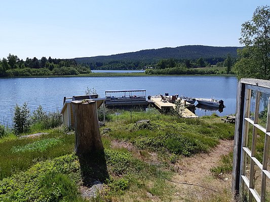 Ein ruhiger See mit einem Steg, an dem mehrere Boote festgemacht sind, umgeben von grüner Vegetation und einem bewaldeten Ufer im Hintergrund.