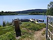 Ein ruhiger See mit einem Steg, an dem mehrere Boote festgemacht sind, umgeben von grüner Vegetation und einem bewaldeten Ufer im Hintergrund.