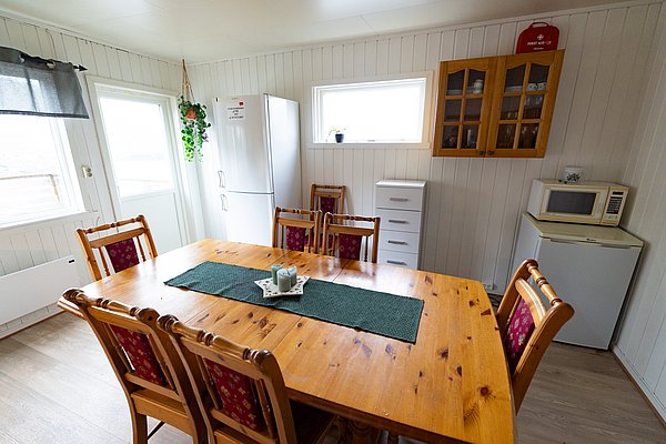 Gemütliche Küche in Nordland, Norwegen: Hölzerner Esstisch mit acht Stühlen, rustikale Einrichtung. Kühlschrank, Mikrowelle und Fenster sorgen für helles, einladendes Ambiente. Ideal für entspannte Angelabende mit Freunden.