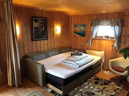 Gemütliche Holzhütte auf Hitra, Norwegen: Wohnzimmer mit ausziehbarem Sofa, Wandbildern und Fensterblick. Ideal für Angelreisen und erholsamen Angelurlaub. Willkommen in der entspannten Atmosphäre dieser charmanten Unterkunft.