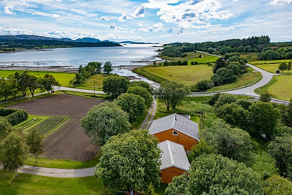 Luftaufnahme von Hitra, Norwegen: Grüne Felder, Küstenlinie und ruhiges Wasser treffen auf ein Holzhaus. Idyllische Landschaft, ideal für Angelurlaub und Naturgenuss. Klarer Himmel schafft eine einladende Atmosphäre für Outdoor-Abenteuer.