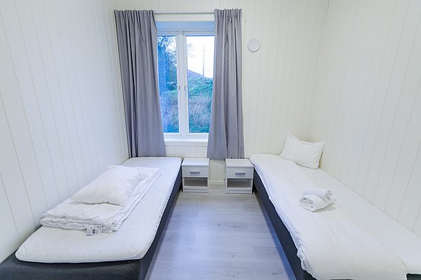 Kompaktes Schlafzimmer mit zwei Einzelbetten, schlichter Dekor und großem Fenster mit Blick auf Norwegens Natur. Ideal für Erholung nach einem aufregenden Angeltag in Nordland, Norwegen. Helle Wände, minimalistisches Design, komfortable Atmosphäre.