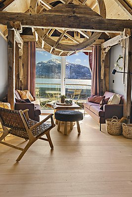 Gemütliches Wohnzimmer mit rustikalen Holzbalken in Südwest-Norwegen. Panoramablick auf verschneite Berge und funkelndes Wasser durch große Fenster. Idealer Rückzugsort für Naturliebhaber und Angel-Enthusiasten. Perfekte Mischung aus Komfort und skandinavischem Charme.