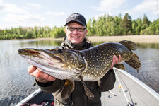 Ein Angler hält stolz einen gefangenen Hecht vor einer malerischen Kulisse aus Wasser und bewaldetem Ufer.