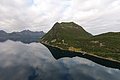 Küste Nord-Norwegens mit markanten Bergen und spiegelglattem Fjord unter bedecktem Himmel. Üppige grüne Landschaft wirkt beruhigend. Ideal für Angelurlaub und Naturerkundung. Abenteuer und Entspannung in beeindruckender nordischer Kulisse.