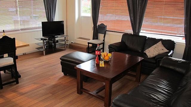 Gemütliches Wohnzimmer in Hitra, Norwegen: Dunkle Ledersofas, Holztisch mit Obstschale, Fernseher auf Regal, großes Fenster mit Jalousien. Idealer Rückzugsort nach einem erfolgreichen Angeltag in der beeindruckenden norwegischen Natur.