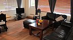 Gemütliches Wohnzimmer in Hitra, Norwegen: Dunkle Ledersofas, Holztisch mit Obstschale, Fernseher auf Regal, großes Fenster mit Jalousien. Idealer Rückzugsort nach einem erfolgreichen Angeltag in der beeindruckenden norwegischen Natur.