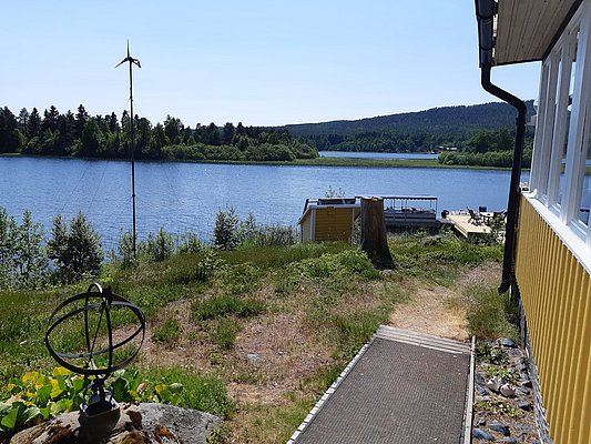 Ein gelbes Haus mit einem Ausblick auf einen ruhigen, von Bäumen umgebenen See unter klarem Himmel, mit einem kleinen Windrad und einer Terrasse im Vordergrund.