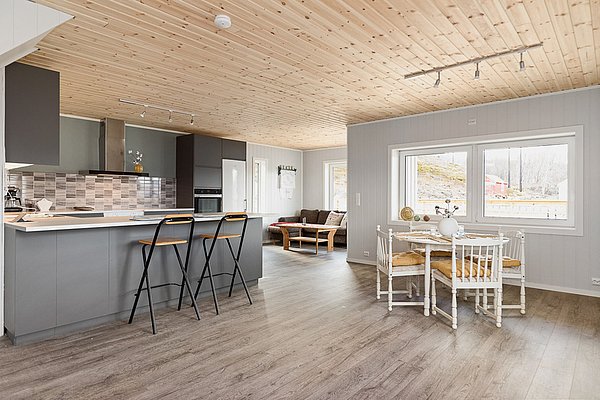 Gemütlicher, moderner Wohnraum in Nordland, Norwegen. Offene Küche mit Barhockern, heller Essbereich und Sofa. Große Fenster bieten Blick auf nordische Natur. Ideal für erholsamen Angelurlaub in skandinavischer Umgebung.