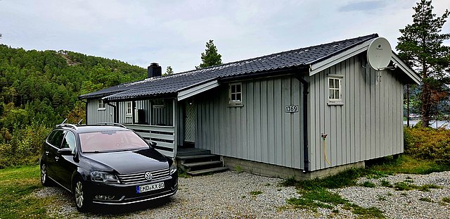 Graues Holzhaus in Norwegens Hitra, umgeben von Waldlandschaft, bietet ideale Unterkunft für Angelurlaub. Im Vordergrund parkt ein Auto; perfekte Basis für Entspannung und Abenteuer in der Natur.