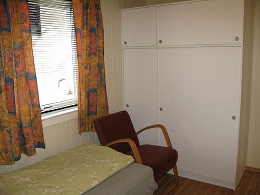 Gemütliches Zimmer in Nordland, Norwegen mit Einzelbett, rotem Sessel, kleinem Fenster und weißen Einbauschränken. Bunte Vorhänge und Holzfußboden schaffen eine warme, einladende Atmosphäre - ideal für entspannte Angelurlaube.