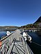 Auf dem Bild ist ein langer Steg in der Region Hitra, Norwegen, zu sehen. Im Hintergrund erstreckt sich bewaldete Landschaft, Boote liegen an der Anlegestelle. Perfekt für Angelbegeisterte und Naturfreunde.