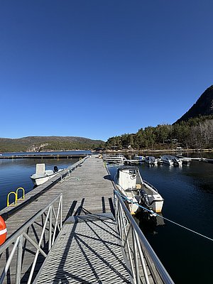 Auf dem Bild ist ein langer Steg in der Region Hitra, Norwegen, zu sehen. Im Hintergrund erstreckt sich bewaldete Landschaft, Boote liegen an der Anlegestelle. Perfekt für Angelbegeisterte und Naturfreunde.