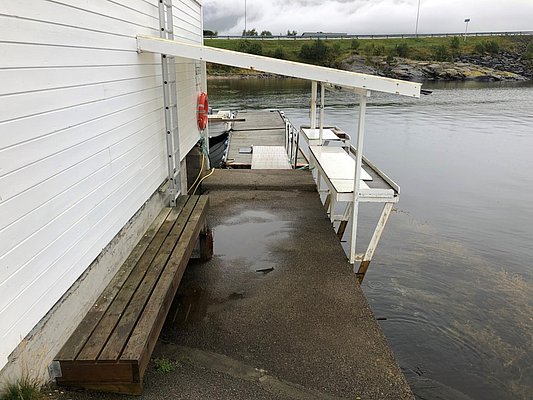 Holzsteg in Hitra, Norwegen: Anglerplattform am Wasser mit Bank, Kutteranlegeplatz und Reinigungsstation. Perfekte Basis für Meeresangeln-Abenteuer. Wolkenverhangen, ruhige Atmosphäre. Anfrage Buchungen für Angelreisen.