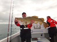 Ein Angler hält einen großen Kabeljau auf einem Boot der "Iceland Sea Angling", während eine weitere Person im Hintergrund zu sehen ist, mit Angelruten an der Reling und bewölktem Himmel im Hintergrund.