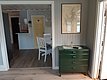 Ein gemütliches, lichtdurchflutetes Zimmer mit Holzboden zeigt einen grünen Schrank unter einer gerahmten Karte, und im Hintergrund sind weiße Möbel und eine gelbe Tür zu sehen.
