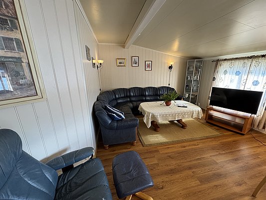 Gemütliches Wohnzimmer in Hitra, Norwegen: Ledersitzgruppe, Holzboden, helle Wände, Tisch mit Tischdecke, Fernseher, Kunstwerke und Pflanzen, ideal für entspannte Angelabende. Perfekter Rückzugsort nach erfolgreichen Meeresangeltouren.