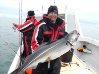 Eine Person in wetterfester Kleidung hält einen großen Fisch an Bord eines Bootes, während eine weitere Person im Hintergrund angelt.