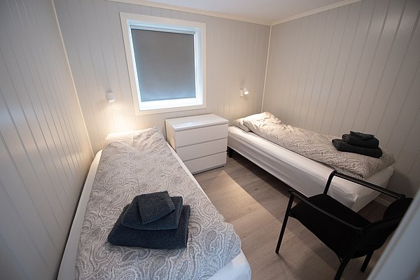 Ein minimalistisches Schlafzimmer mit zwei Einzelbetten, grauer Bettwäsche und einem Fenster zwischen den Betten.