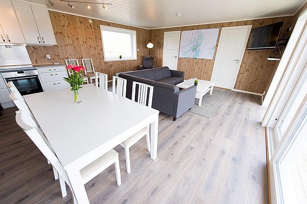 Modernes skandinavisches Ferienhaus in Nordland, Norwegen. Helles Wohnzimmer mit Holzwänden, gemütlicher Sitzecke, Esstisch, Küche und großen Fenstern. Ideal für entspannte Angelurlaube mit Komfort und Stil.
