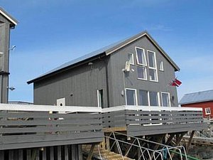Graues, skandinavisches Fischerhaus auf Stelzen in der Region Nordland, Norwegen. Typische Architektur, Holzverkleidung und norwegische Flagge. Ideal für Angelreisen oder Küstenabenteuer in beeindruckender, natürlicher Landschaft. Klarer, blauer Himmel im Hintergrund.