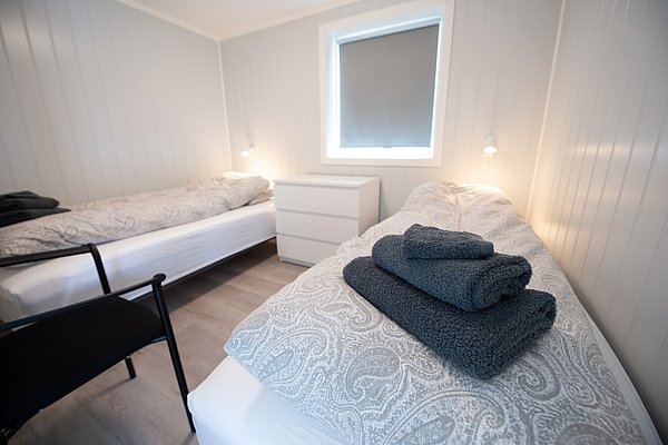 Helles, minimalistisch eingerichtetes Schlafzimmer mit zwei Einzelbetten, grauer Bettwäsche und gefalteten Handtüchern in der Mitte.