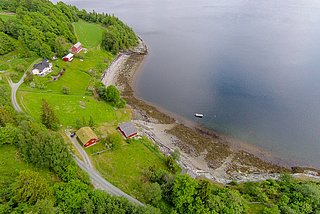 Luftaufnahme einer idyllischen Küstenlandschaft in Nord-Norwegen: grüne Wiesen, rote Holzhäuser und ein ruhiger Fjord. Perfekt für Angelreisen und Naturerlebnisse, bietet diese Region atemberaubende Ausblicke und Erholung pur. Ideal für Angelurlaub in Norwegen.
