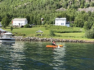 Klares Wasser, idyllische Küstenlandschaft Nord-Norwegens mit traditionellen weißen Häusern und grüner Natur. Ein Kajakfahrer paddelt nahe einem ankernden Boot. Perfekt für Angelreisen und Naturerkundung.