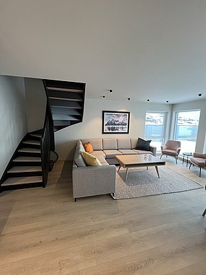 Moderne Ferienwohnung in Nordland, Norwegen: Helle Sitzecke mit gemütlichem Sofa, Holztisch, stilvolle Wendeltreppe, große Fenster mit Blick auf die faszinierende Landschaft. Perfekt für entspannte Angelurlaube und Erholung in skandinavischem Design.