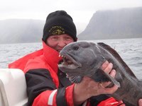 Ein Angler in roter Outdoor-Bekleidung hält einen Wolfsfisch vor einer nebligen, bergigen Küstenlandschaft.
