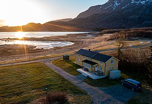 Das Bild zeigt ein gelbes Haus in Nordland, Norwegen, bei Sonnenuntergang. Ein ruhiger Fjord und majestätische Berge im Hintergrund bieten eine idyllische Landschaft. Ideal für ruhige Angelreisen und Naturerlebnisse.