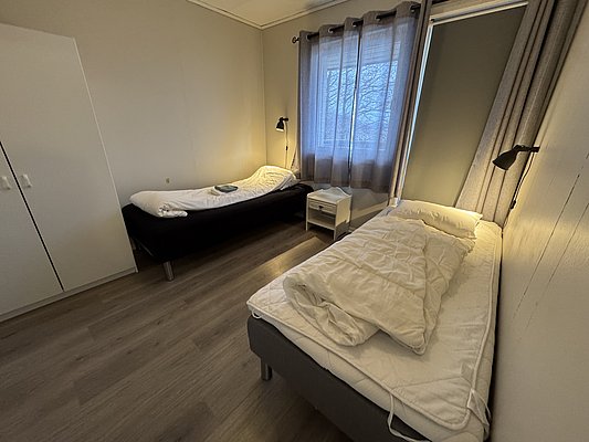 Gemütliches Schlafzimmer mit zwei Einzelbetten in einer Unterkunft in Hitra, Norwegen. Helle Holzböden, Fenster mit Vorhängen, Leselampen neben den Betten. Ideal für Angler auf Angelreisen. Ruhiges, einladendes Ambiente für entspannte Nächte.