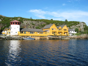 Gelbe Holzhäuser und runder Leuchtturm am Wasser in Südwest-Norwegen, umgeben von felsiger Landschaft und klarem Himmel. Ideal für Angelurlaube und Naturerlebnisse. Ein perfekter Ort für Entspannung und Abenteuer in der Natur.