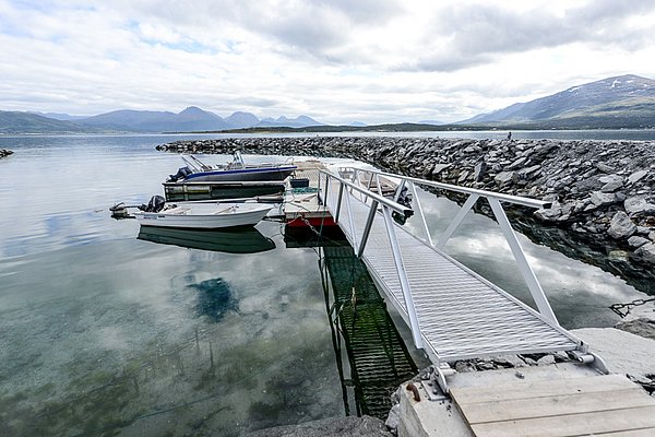 Fjordblick in Nord-Norwegen: Boote am Steg, klare Gewässer, umrahmt von majestätischen Bergen und grauem Himmel. Ideal für Angelreisen und Naturerlebnis.