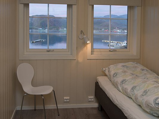 Ein gemütliches Schlafzimmer mit einem Bett und einem Stuhl, mit Blick aus zwei Fenstern auf einen ruhigen See und eine bergige Landschaft im Hintergrund.