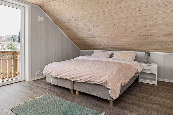 Gemütliches Schlafzimmer in Nordland, Norwegen: Hell gestaltet mit Holzverkleidung, großem Doppelbett, Nachttisch und Lampe. Blick auf Balkon und Waldlandschaft. Ideal für Angelreisen und Entspannung.