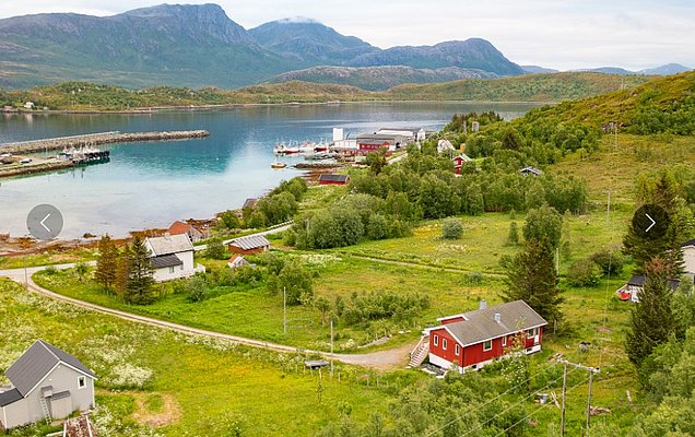 Idyllische Küstenlandschaft Nord-Norwegens mit roten Holzhäusern, sanften Hügeln und einem ruhigen Fjord, umgeben von üppigem Grün und majestätischen Bergen. Ideal für Angelurlaub und Naturliebhaber.