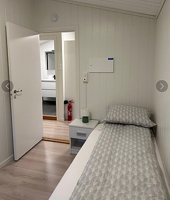 Gemütliches Schlafzimmer mit Einzelbett, modernem Nachttisch und Tür zum Badezimmer. Helle Holzwände und -böden schaffen eine einladende Atmosphäre. Ideale Unterkunft für Angelurlaub in der Region Hitra, Norwegen.