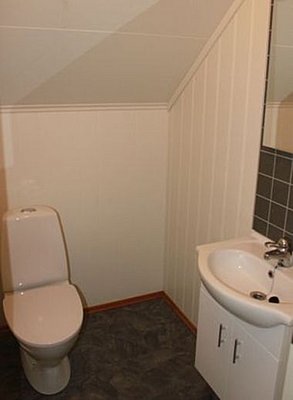 Kompaktes, schlichtes Badezimmer in Nord-Norwegen: weiße Wände, Dachschräge, grauer Boden, Keramiktoilette, kleines Waschbecken mit Spiegel, ideal für entspannte Angelreisen-Unterkunft. Perfekt für komfortable Erholung nach einem fischreichen Tag.