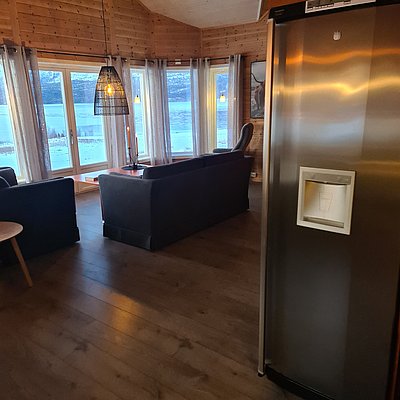 Gemütliches Wohnzimmer in Nord-Norwegen mit Holzwänden und großen Fenstern, die winterliche Landschaft zeigen. Gemütliche Sitzecke, ein moderner Kühlschrank und gedimmtes Licht schaffen eine warme, einladende Atmosphäre. Ideal für Angelreisen und Entspannung.