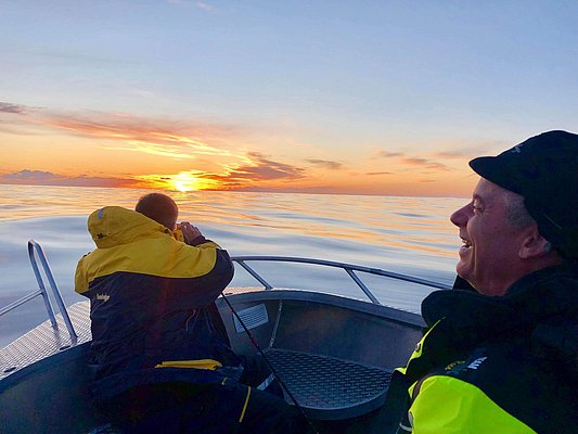 Zwei Angler genießen einen atemberaubenden Sonnenuntergang auf einem Boot in Nordland, Norwegen. Der Himmel spiegelt sich im ruhigen Meer und schafft eine idyllische Atmosphäre für einen unvergesslichen Angelurlaub.
