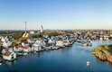 Luftaufnahme eines malerischen Küstendorfs mit weißen Häusern, einem Leuchtturm und mehreren Booten im ruhigen blauen Wasser. Luftaufnahme eines malerischen Küstendorfs mit weißen Häusern, einem Leuchtturm und mehreren Booten im ruhigen blauen Wasser.