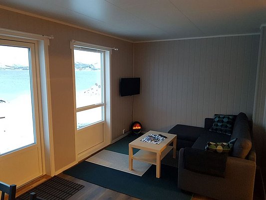 Gemütliches Wohnzimmer in Nord-Norwegen mit modernem Sofa, Tisch und dekorativem Kamin. Große Fenster bieten Blick auf verschneite Landschaft und Meer. Perfekte Unterkunft für erholsamen Angelurlaub und Naturgenuss.