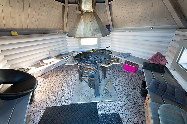 Gemütliche Grillhütte in Nord-Norwegen mit zentralem Grillplatz, Holzbänken und Kissen. Perfekt zum Angeln, Entspannen und für gesellige Abende nach erfolgreichen Angelstunden in der arktischen Natur.