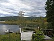 Ein Holzdeck mit Blick auf einen ruhigen See, umgeben von üppigem Grün und Hügeln, unter einem bewölkten Himmel.