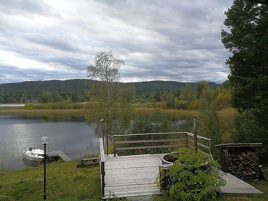 Ein Holzdeck mit Blick auf einen ruhigen See, umgeben von üppigem Grün und Hügeln, unter einem bewölkten Himmel.