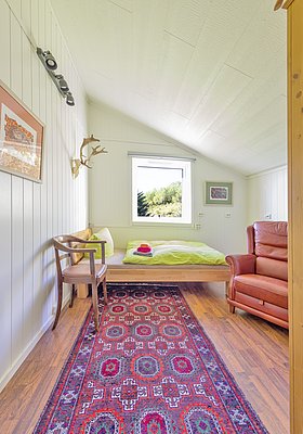Gemütliches Zimmer in Nordland, Norwegen: Holzboden, bunte Wanddekorationen und ein Fenster mit Ausblick auf grüne Wälder. Ideal für erholsame Angelurlaube. Harmonisches Ambiente mit rotem Teppich, Bett und Sessel.