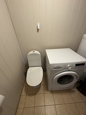 Waschraum in Hitra, Norwegen: Moderne Waschmaschine neben einer Toilette auf hellen Fliesen. Funktionale Ausstattung für Reisende, die Komfort und Praktikabilität in ihrer Unterkunft suchen. Ideal für Angelreisende in Norwegen.